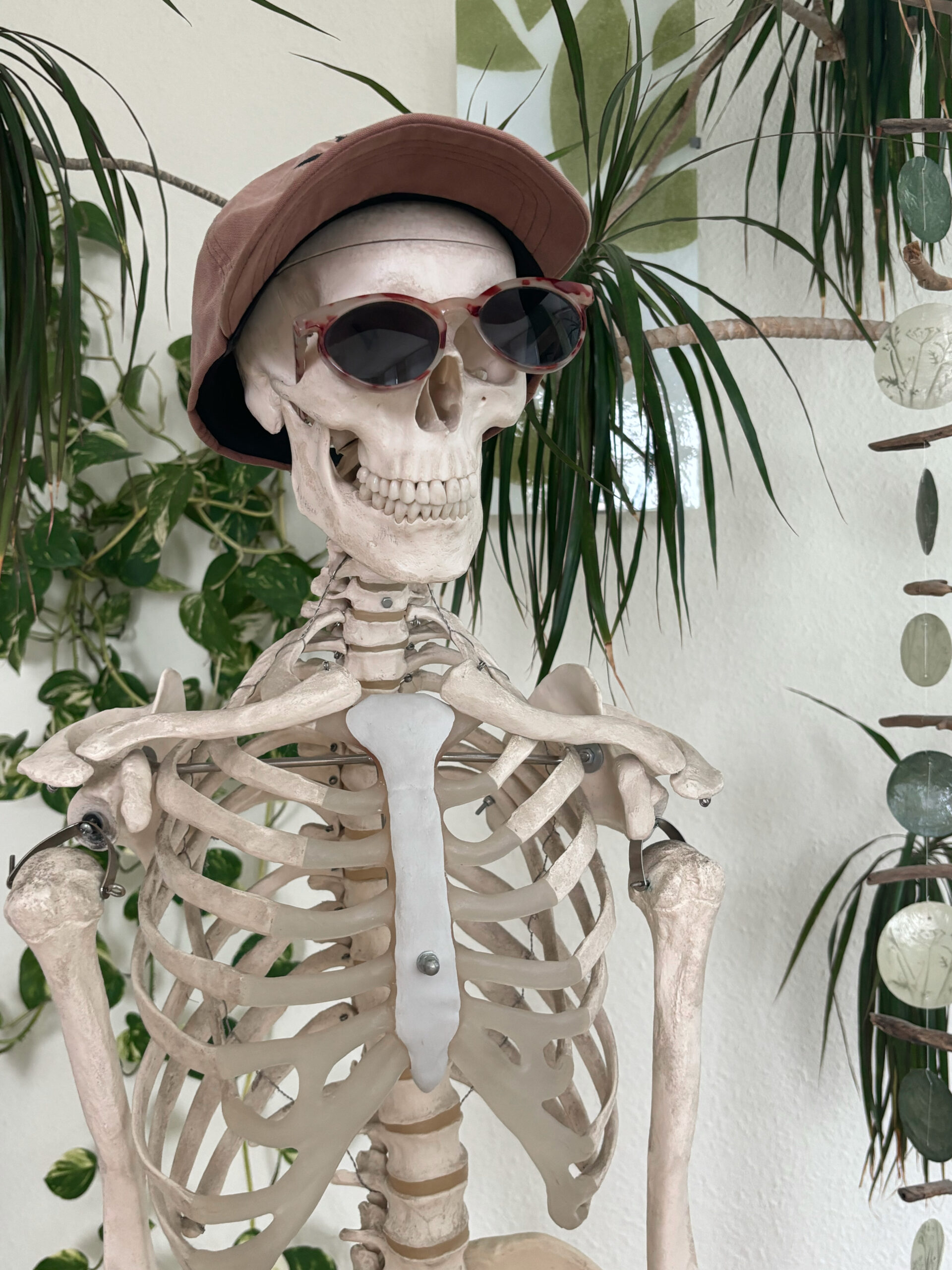 Ein freundliches Skelett mit Sonnenbrille und Kappe als Nahaufnahme mit vielen Blumen im Hintergrund bei Physio Ganster